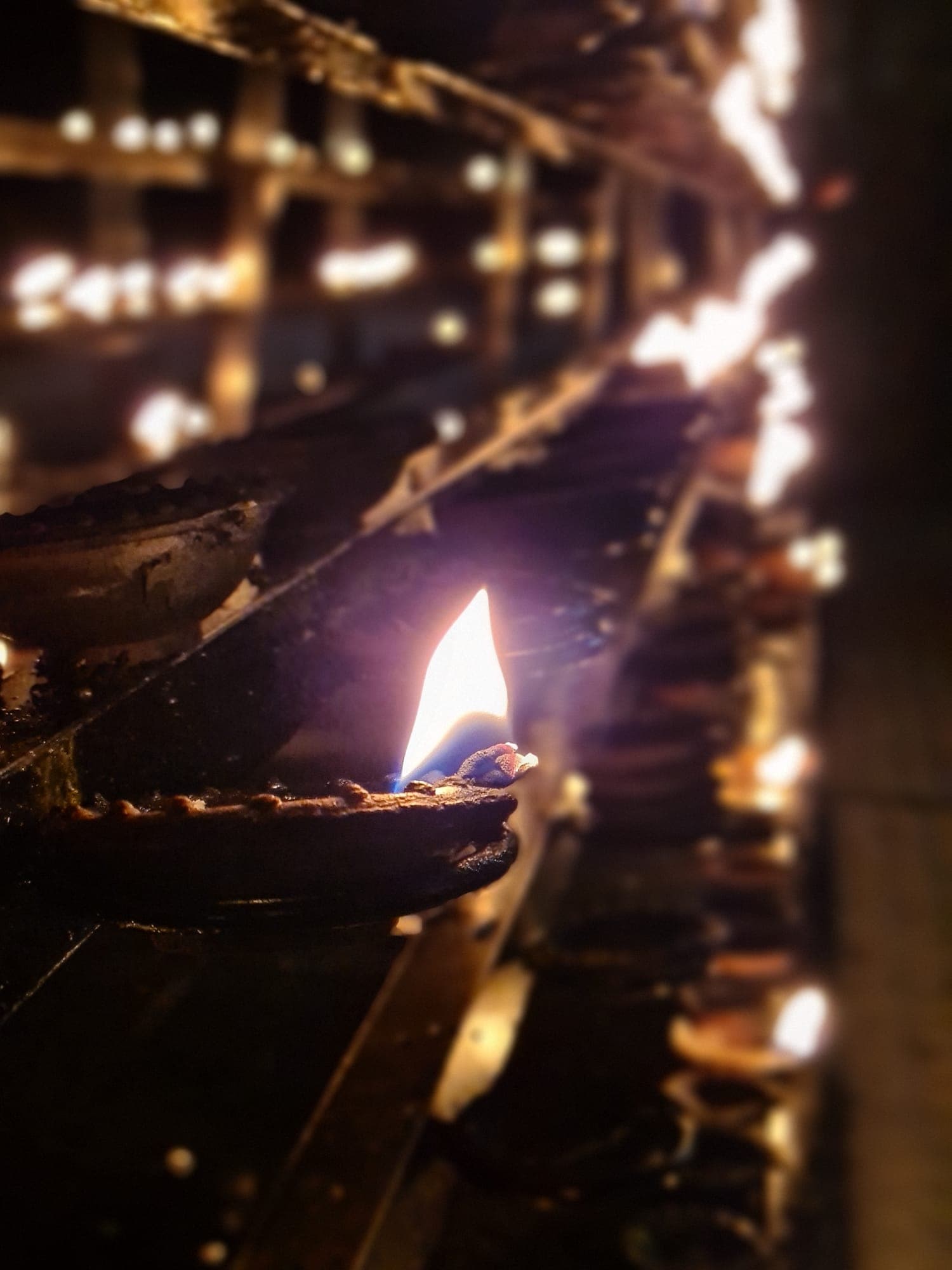 A single oil-lamp flame in sharp focus, with rows of lit clay lamps glowing softly behind it.