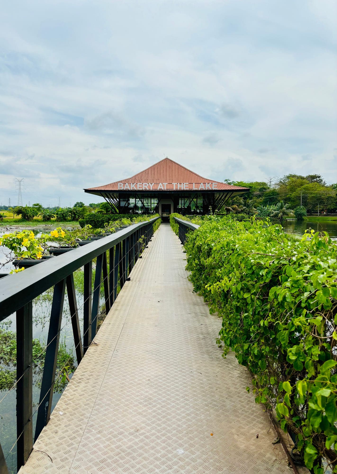 A long walkway with railings and flowering plants leading across a lake to a pavilion with a sign reading 'Bakery at the Lake'.