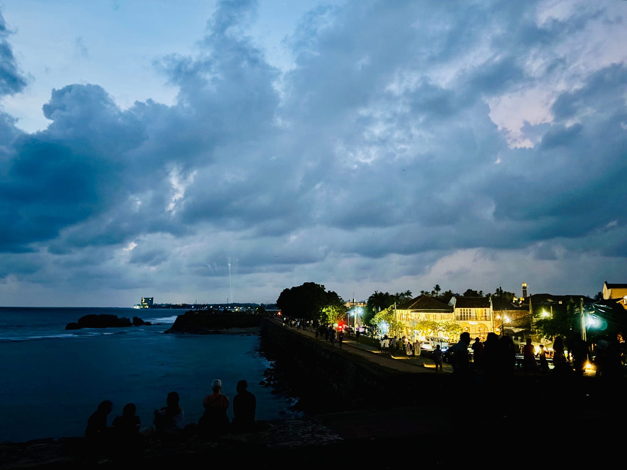 People sitting on the Galle Fort rampart at dusk under heavy clouds, with the sea on the left and lit colonial buildings along the wall.