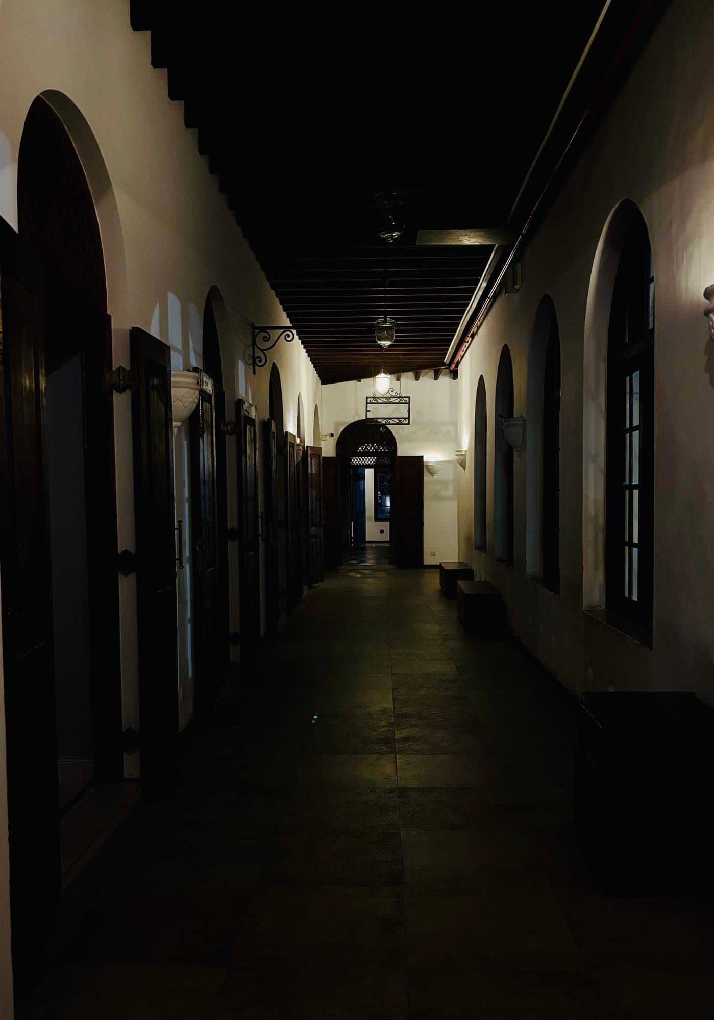 A long, dimly lit colonial corridor with arched windows and doors on both sides, ending in an arched doorway.