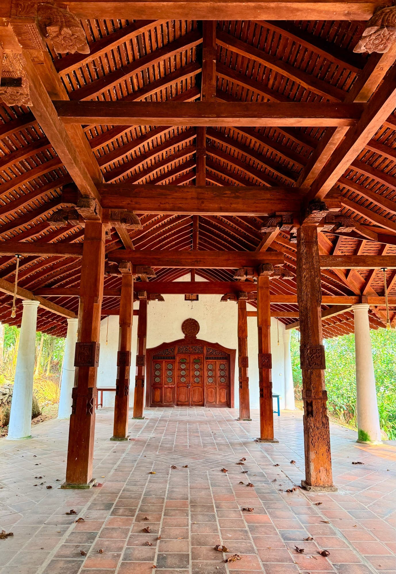A traditional open pavilion with carved wooden pillars and a timber roof over a brick floor, leading to ornate wooden doors.