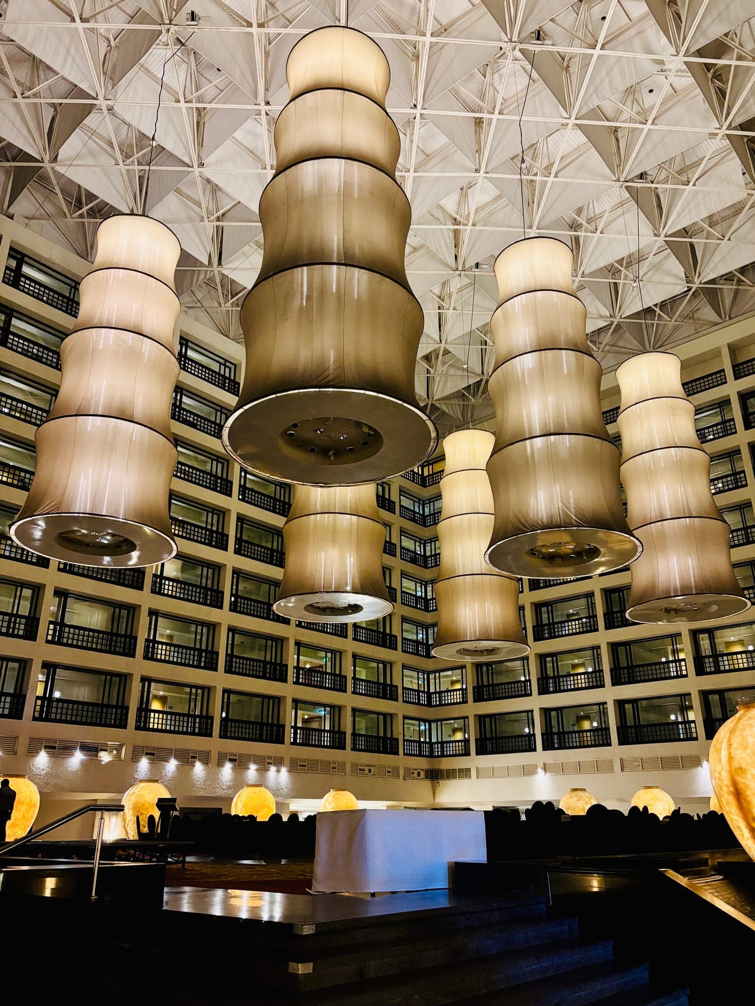Tall layered fabric lanterns hanging in a hotel atrium beneath a white geometric ceiling, with rows of balconies behind.