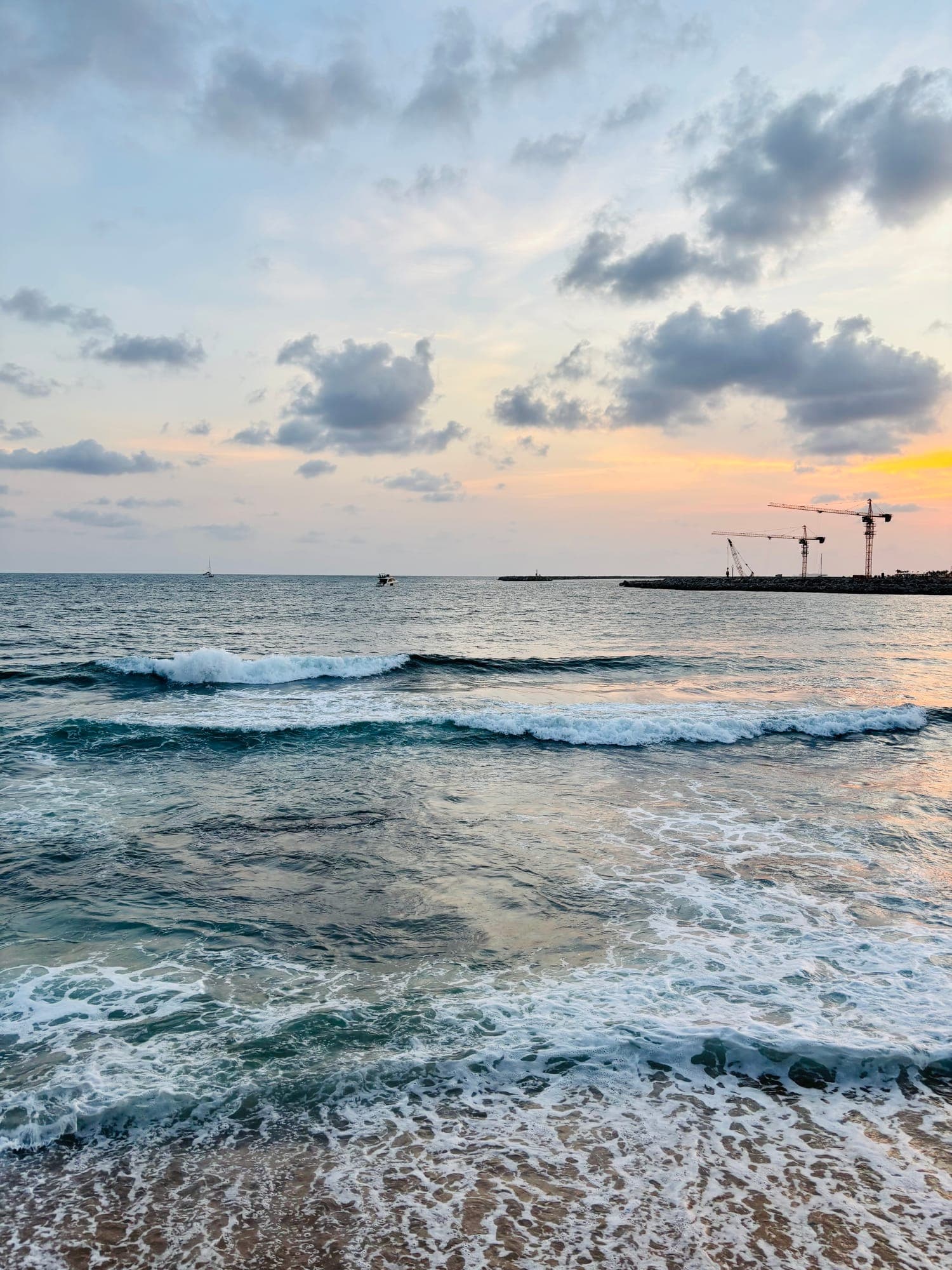 Waves rolling onto the shore at sunset, with construction cranes on a breakwater in the distance.