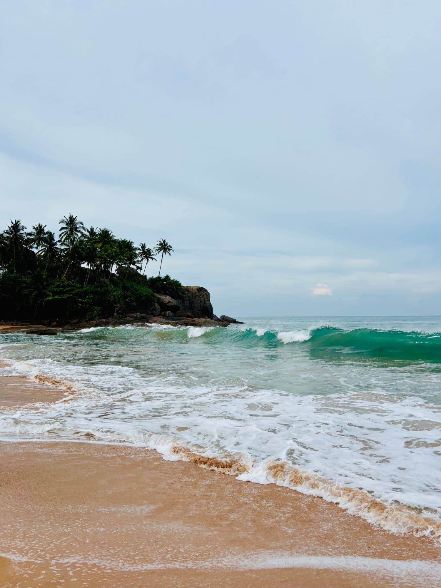 Turquoise waves breaking on a sandy beach beside a rocky headland covered in palm trees, under an overcast sky.