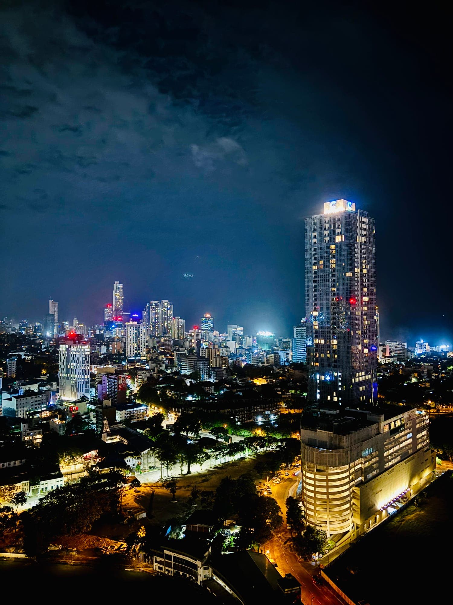 A city skyline at night under a cloudy sky, with a tall lit tower in the foreground and streets glowing below.
