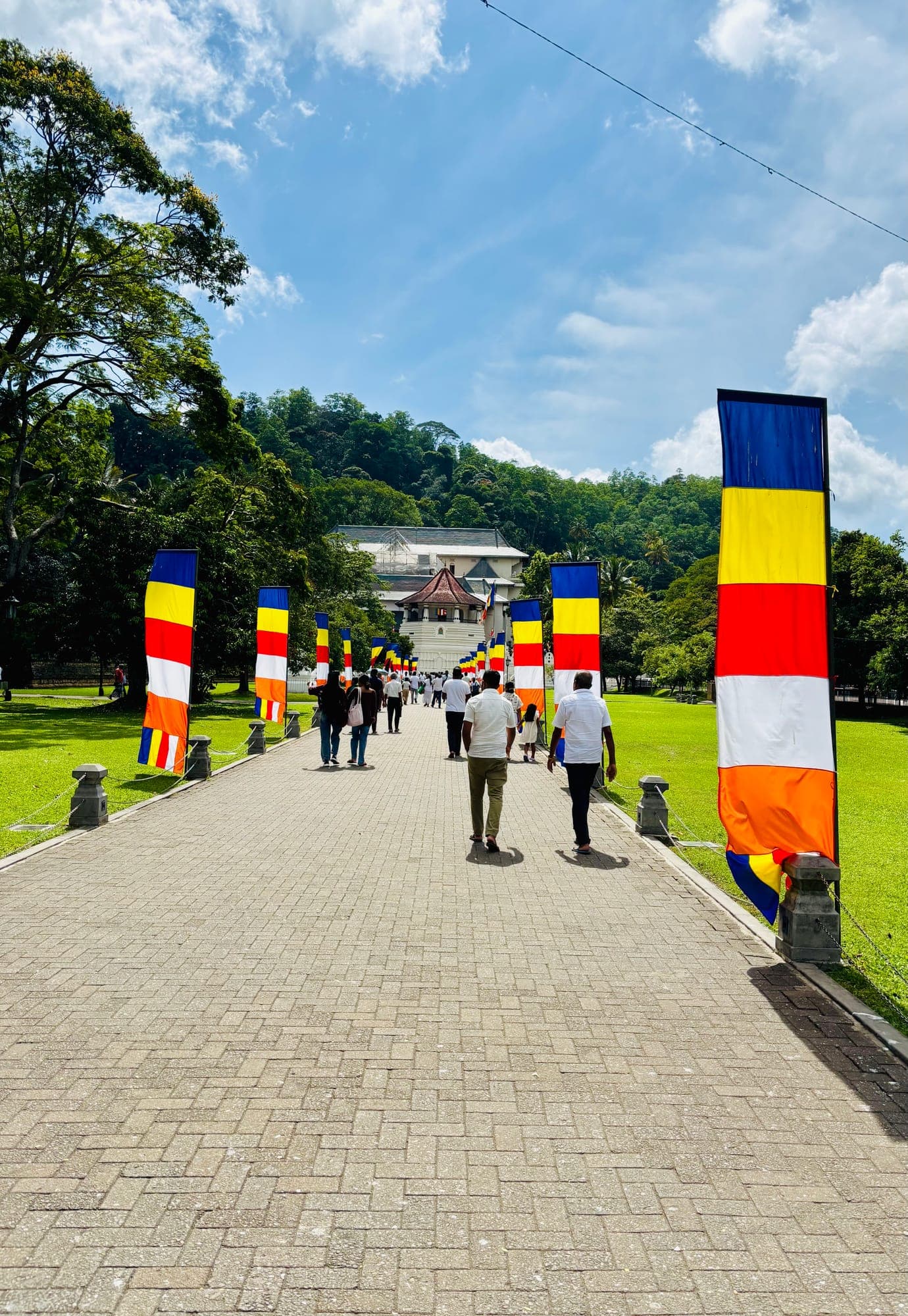 A paved walkway lined with tall Buddhist flags leading toward the Temple of the Tooth, with visitors walking under a bright sky.