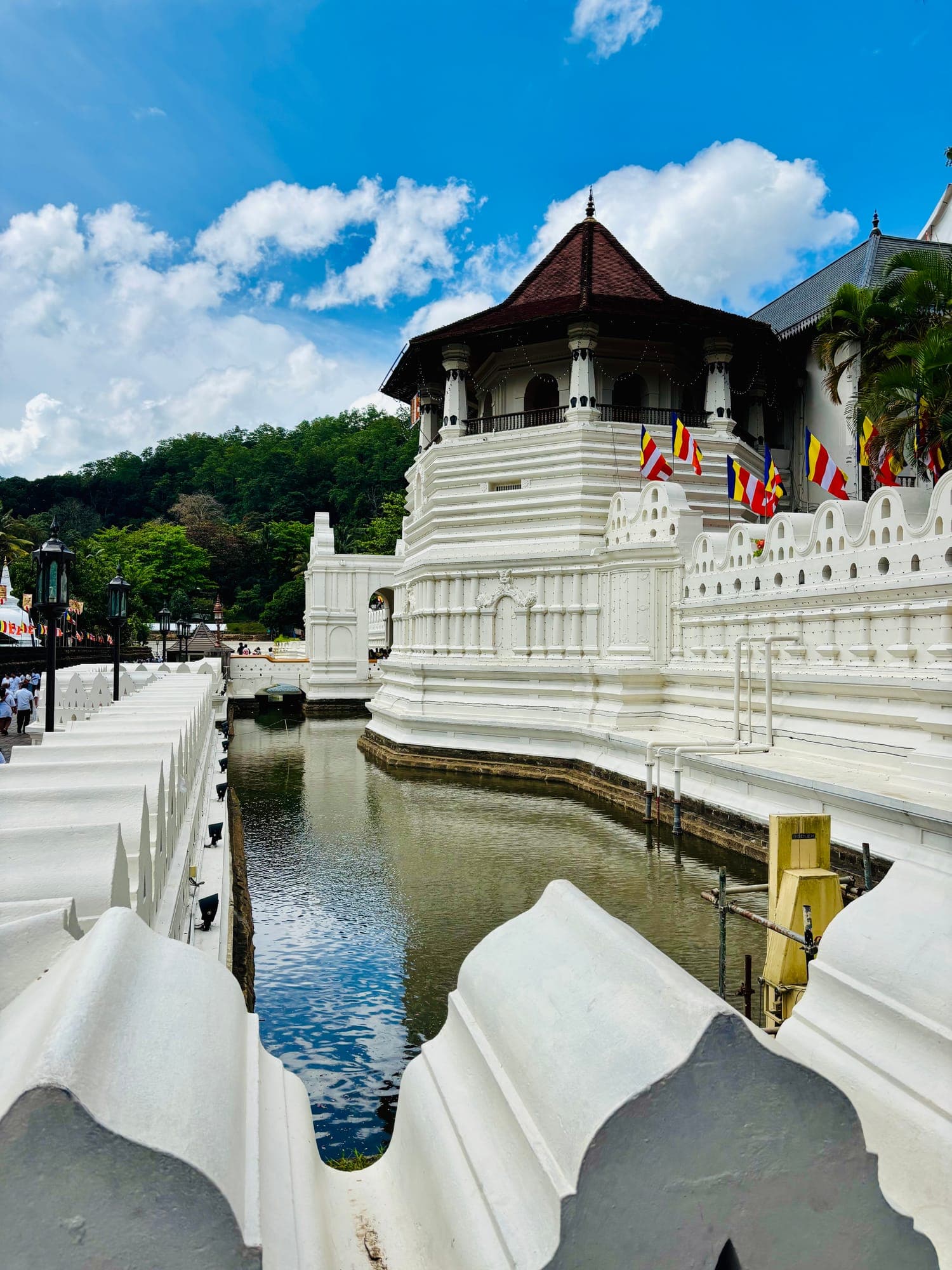 The white octagonal pavilion of the Temple of the Sacred Tooth Relic in Kandy, with Buddhist flags, reflected in the moat under a blue sky.